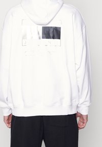 Felpa bianca con cappuccio caratterizzata da una grande grafica sul retro con sezioni rettangolari nere e argentate, testo "TOMMY JEANS" e polsini a coste.