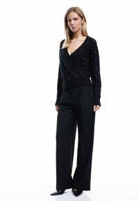 Schwarzer Wickelstrickpullover mit V-Ausschnitt und Perlenakzenten, kombiniert mit hochtaillierten schwarzen Hosen und spitzen schwarzen Absätzen. Glatte Textur und taillierte Passform.