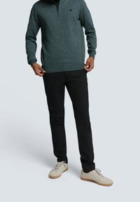 Homme portant un pull à col zippé vert foncé, un pantalon noir et des baskets beiges à semelles gomme, debout sur un fond gris clair.
