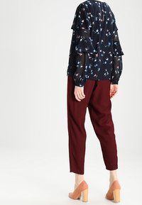 Blusa floreale scura con maniche a ruffle sovrapposte, abbinata a pantaloni larghi borgogna e scarpe con tacco a blocco marrone chiaro.