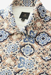 Præget skjorte med blomsterdesign i beige, blå og brun; har kravesnit og runde knapper. Etiket angiver mærket "NEXT".