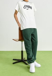 Vit t-shirt med svart "Nike"-logotyp, i kombination med mörkgröna sportbyxor och vita sneakers. Modellen står bredvid en trärstol.