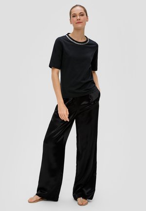 Zwarte top met korte mouwen en een metallic-accent bij de hals, gecombineerd met zwarte satijnen broek met wijde pijpen en zilveren teenslippers. Glad textuur.