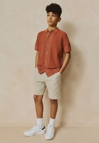 Camicia a maniche corte di colore ruggine con cuciture bianche, abbinata a pantaloncini beige chiaro e sneakers bianche con dettagli blu navy.