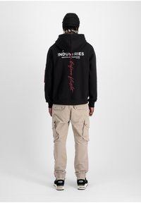 Sudadera negra con texto gráfico en blanco y rojo, combinada con pantalones cargo beige que tienen múltiples bolsillos. Zapatillas negras completan el conjunto.