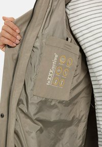 Die wasserfeste beige Jacke hat ein glattes Innenfutter, eine Tasche mit Pflegeinformationen und Symbole, die auf die wasserfesten und atmungsaktiven Eigenschaften hinweisen.