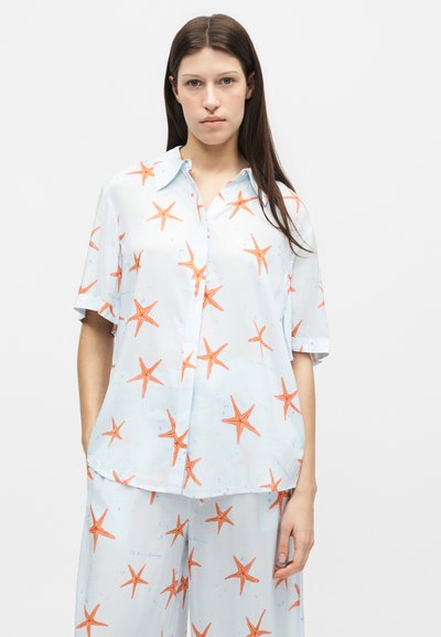Femme portant une chemise bleu clair à manches courtes et un pantalon avec imprimé étoiles de mer orange, debout devant un fond blanc uni.