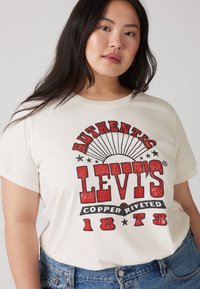Kreminė marškinė, kuriose yra "AUTHENTIC LEVI'S" raudona ir juoda spalvomis, su senovišku logotipu ir dizaino elementais, tokiais kaip žvaigždės ir saulės spinduliai. Matosi mėlynos džinsinės kelnes.