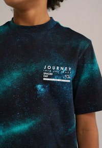Námořnické tričko s krátkým rukávem, které má galaxii vzor v modrých a zelených odstínech, s bílým grafickým textem a akcenty na přední straně.
