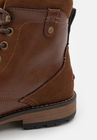 Brązowa skórzana i zamszowa wysoka boot hiking z okrągłymi sznurówkami, oczkami w kolorze złota, widocznymi szwami oraz teksturowaną gumową podeszwą.