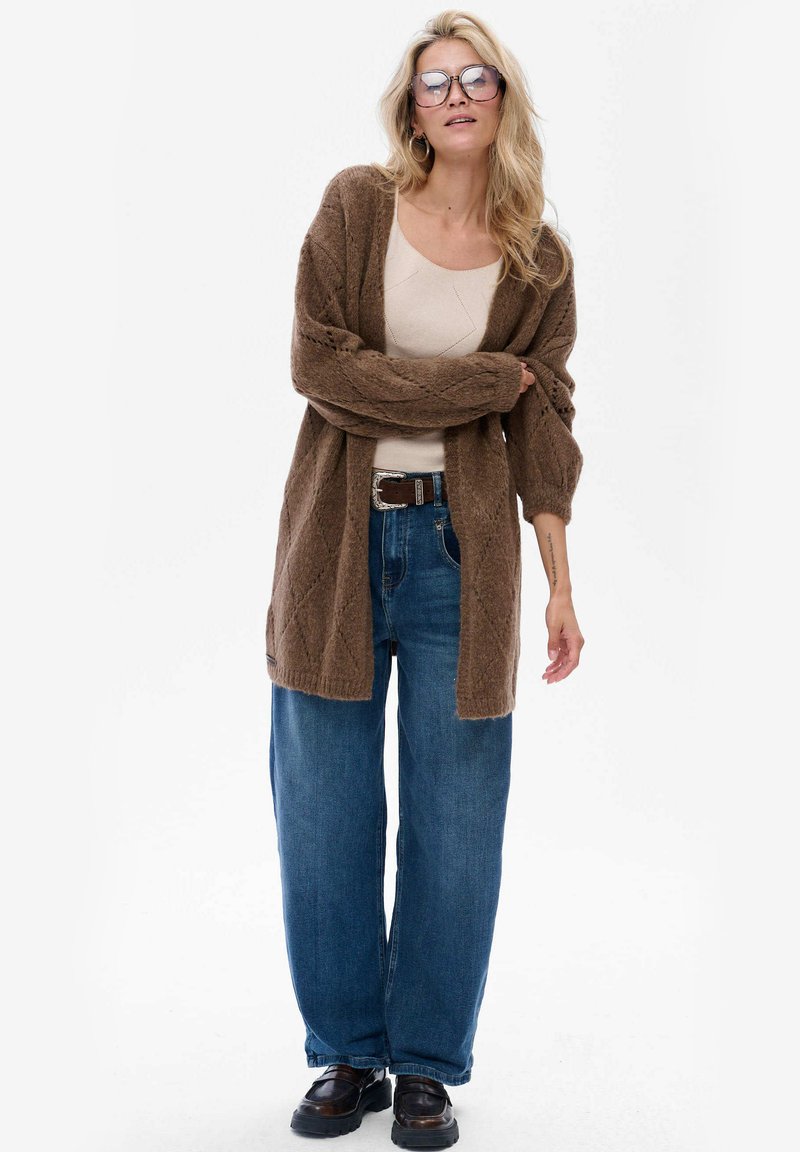 Bruine gebreide cardigan met een textuurpatroon, gedragen over een beige top. Gecombineerd met losse blauwe spijkerbroeken en zwarte schoenen. Draagt een paar duidelijke bril.