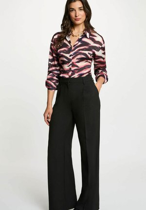 Mujer con blusa de manga larga de estampado rosa y negro, metida dentro de pantalones negros de pierna ancha y cintura alta, de pie con una mano en el bolsillo.