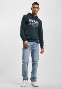 Dark green hoodie with logo, kangaroo pocket, and ribbed cuffs, paired with light blue jeans and white sneakers. Simple and casual design.