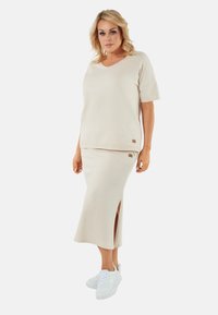 Beżowa bluzka z dekoltem w serek i krótkimi rękawami zestawiona z pasującą długą spódnicą z bocznym rozcięciem, noszona z białymi trampkami.