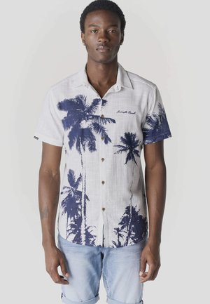 Hombre vistiendo camisa blanca de manga corta con estampado de palmeras azul marino y pantalones cortos de mezclilla azul claro, de pie contra un fondo liso.