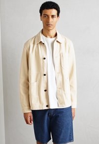 Veste utilitaire beige avec un bouton sur le devant, poche poitrine et poches latérales. Portée par-dessus un T-shirt blanc et assortie à un short en denim bleu.