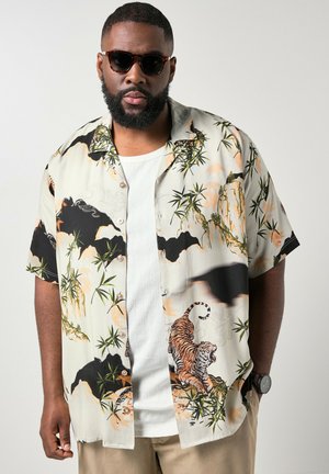 Man die een donkere zonnebril en een beige overhemd met tijger- en bamboeprints draagt over een wit geribd shirt, met kort haar en baard.