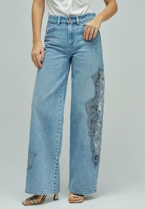 Femme portant un jean large bleu clair avec des patchs en dentelle florale sombre sur les jambes et des sandales à talons ouvertes beige, top blanc court.