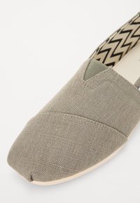 Chaussure à enfiler en tissu gris avec un bout arrondi et une surface texturée. L'intérieur présente un motif chevron en beige et noir.