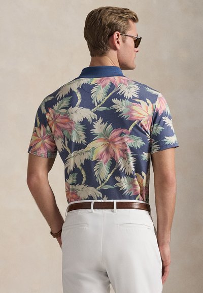 Άνδρας με γυαλιά ηλίου, μπλε πουκάμισο με λουλούδια, λευκό παντελόνι και καφέ ζώνη, στέκεται με την πλάτη γυρισμένη σε ουδέτερο φόντο.