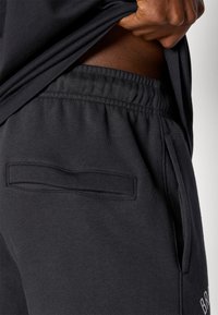 Melni treniņbikses ar elastīgu jostasvietu, plakanu aizmugures kabatu un gludu faktūru, ko izceļ diskrēta logo detaļa sānos.