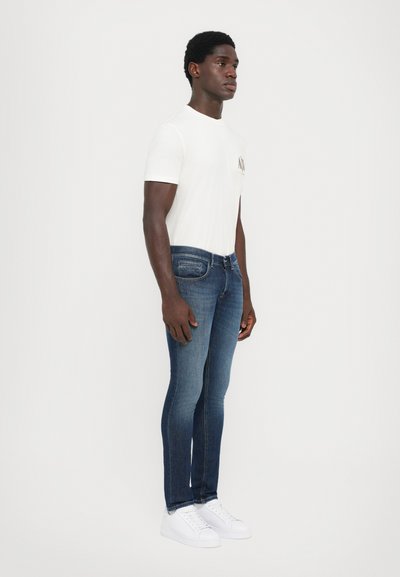 Slim-fit blå jeans med medelhavstvätt och subtila slitage, kombinerat med en vit t-shirt och vita sneakers, på en modell som står sidledes.