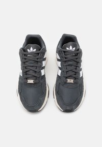 Grå sportskor med svarta och vita ränder, nät- och mockatexturer, rund tå och en uttalad sula. Har Adidas-logotypen.