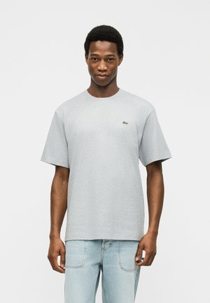 Mladi moški, oblečen v preprosto svetlo sivo kratko majico in svetlo modre kavbojke, stoji pred preprostim belim ozadjem.