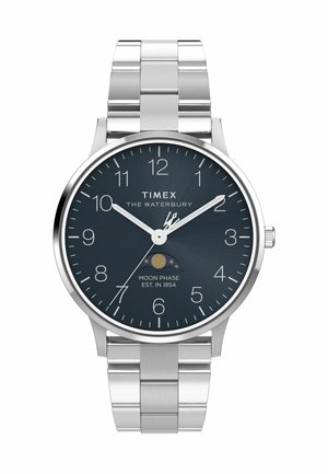 Montre Timex Waterbury avec bracelet en acier inoxydable argenté, cadran bleu marine, index blancs et indicateur de phase lunaire. Design de boîtier circulaire.