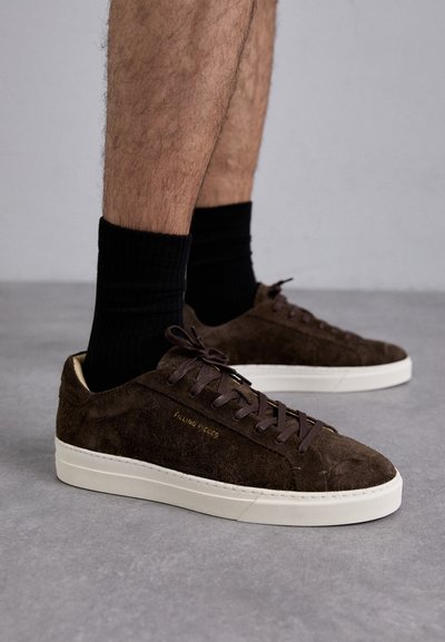 Brown suede sneakers with a white rubber sole, featuring brown laces and subtle branding on the side, paired with black ribbed socks.
