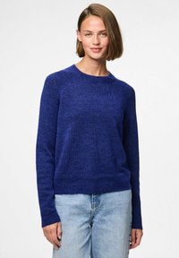 Zils adīts džemperis ar apaļu dekoltē un garām piedurknēm, ar nedaudz brīvu piegriezumu un teksturētu apdari. Kombinēts ar gaišiem džinsa biksēm.