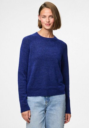 Pull en tricot bleu avec un col rond et des manches longues, présentant une coupe légèrement ample et une finition texturée. Associé à un jean en denim clair.