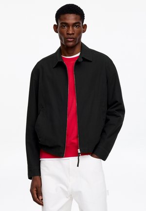 Homme portant une veste noire à fermeture éclair par-dessus un sweat-shirt rouge et un pantalon blanc, debout devant un fond blanc uni.