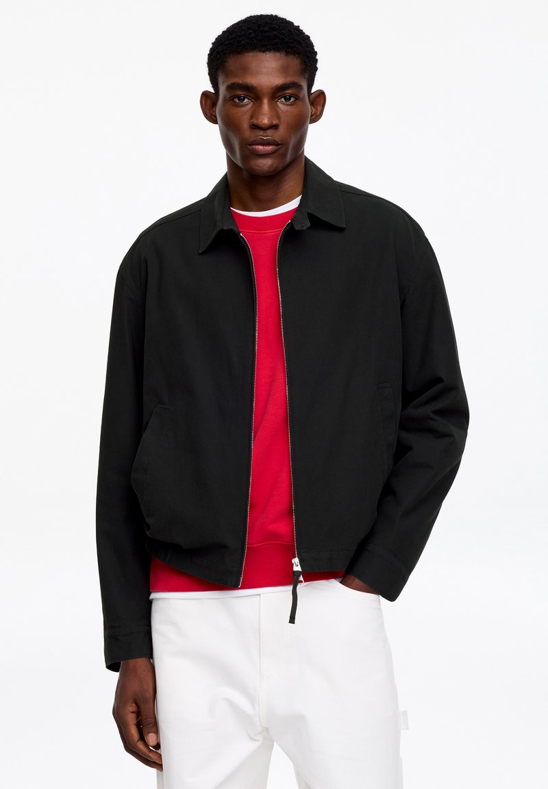 Homme portant une veste noire à fermeture éclair par-dessus un sweat-shirt rouge et un pantalon blanc, debout devant un fond blanc uni.