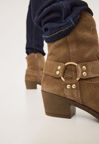Botines de ante marrón con un tacón grueso, adornados con un acento circular dorado y detalles de costura, combinados con denim oscuro.