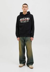 Sudadera negra con un estampado gráfico que presenta texto en blanco y rojo. Unos jeans de mezclilla verde de corte holgado y zapatillas beige completan el atuendo.