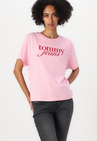 Rosa bomulls T-skjorte med løs passform, med den røde logoen "tommy jeans" i en kursiv skrift. Kombinert med mørke jeans.