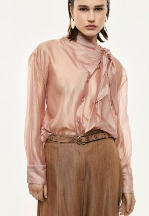 Donna con una camicetta rosa trasparente con un grande fiocco annodato al collo, abbinata a pantaloni marroni a vita alta e una cintura con trama.