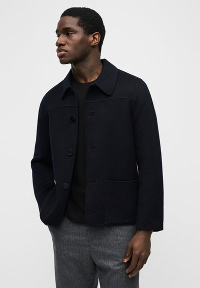 Άνδρας που φοράει μαύρο σακάκι με κουμπιά και γιακά, μαύρο πουκάμισο και γκρι παντελόνι, κοιτάζει προς τα αριστερά με τα χέρια μερικώς στις τσέπες.