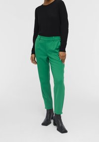 Personne portant un pantalon tailleur vert, un haut à manches longues noir et des bottines à talons carrés noires, se tenant sur un fond blanc.