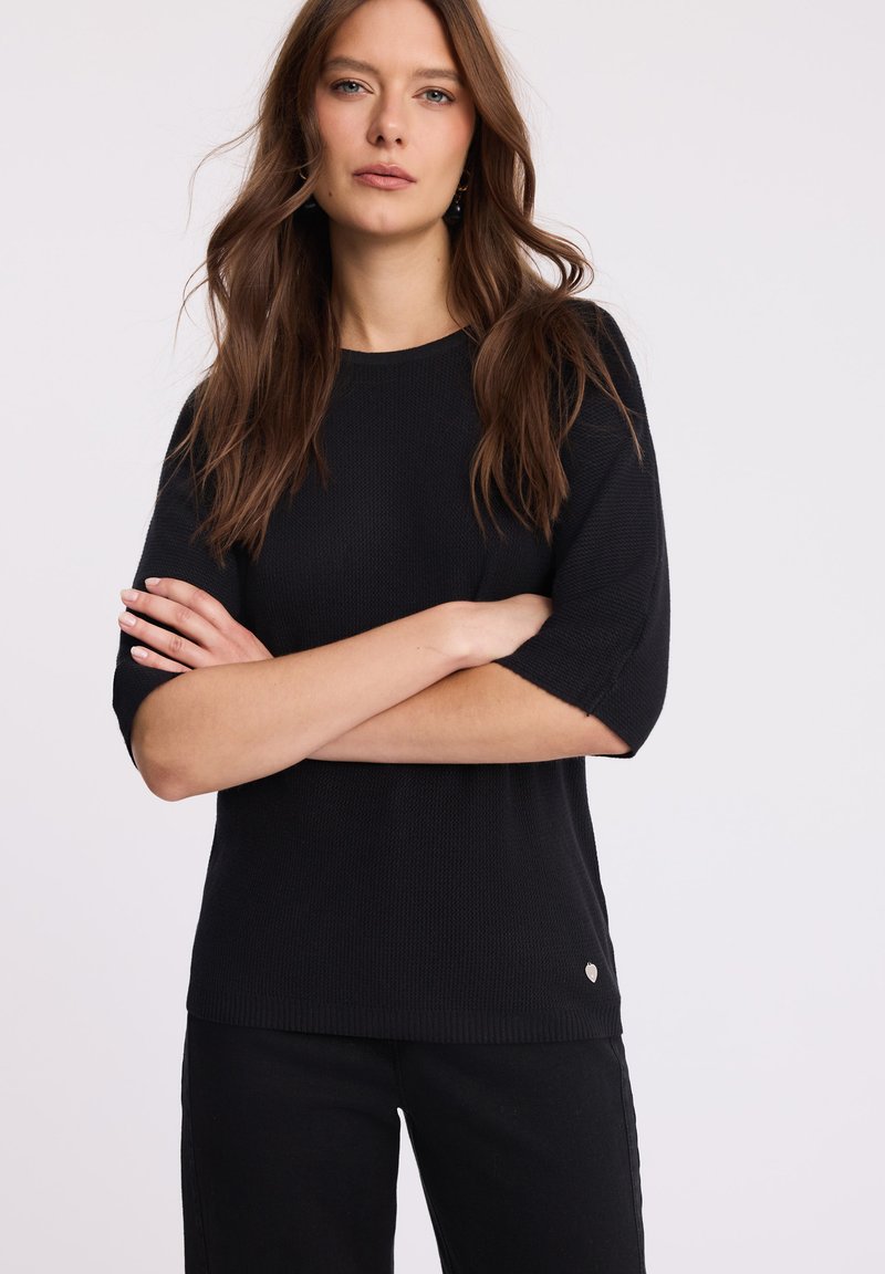 Kobieta z długimi brązowymi włosami w czarnej krótko rękawowej dzianinowej bluzce i czarnych spodniach, z założonymi rękami, pozująca na białym tle.