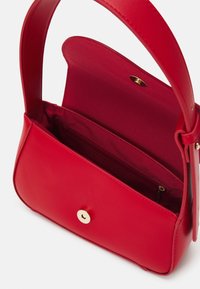 Sac à main en cuir rouge avec rabat ouvert, montrant une poche intérieure zippée et une fermeture bouton magnétique doré, ainsi qu'une bandoulière simple.