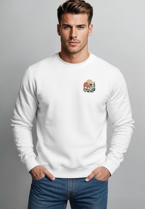 Young man with styled hair wears white sweatshirt with mountain, tree, river graphic on chest, blue jeans, hands in pockets, neutral background.