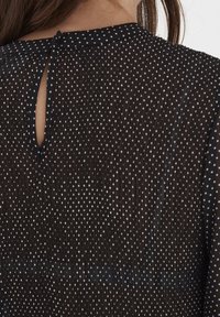 Blusa nera trasparente con pois bianchi, caratterizzata da una chiusura a occhio di bue sul retro e pieghe testurizzate. Tessuto morbido con un design fluido.