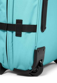 Maleta turquesa con exterior de tela duradera, carcasa de ruedas negra y correas negras texturizadas para mayor soporte.