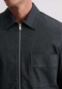 Ciemnoszara kurtka dżinsowa z zamkiem z przodu, spiczastym kołnierzem i naszywaną kieszenią na piersi. Materiał ma miękką fakturę z delikatnym połyskiem.