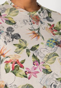 T-shirt med färggrad blommönster och svaga svartvita skisser av tecknade figurer på beige tyg.