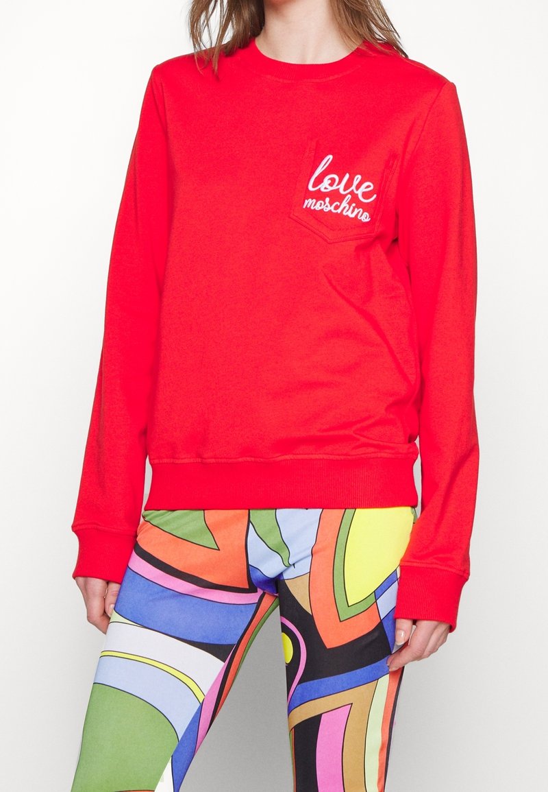 Sudadera roja de manga larga, con cuello redondo y un bolsillo en el pecho. Presenta el texto "love moschino" bordado en blanco. Textura suave y ajuste casual.