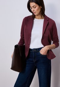 Femme portant un blazer bordeaux, une chemise blanche et un jean foncé, tenant un grand sac fourre-tout marron foncé d'une main, l'autre main dans la poche.