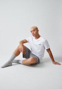 Weißes Baumwoll-T-Shirt mit kleinem schwarzem Logo, kombiniert mit grauen Shorts und hellgrauen gerippten Socken, Modell sitzt vor einem schlichten Hintergrund.